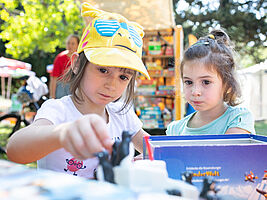 Zu sehen sind zwei Kinder auf einer Spieleveranstaltung im Park. Sie spielen das Geschicklichkeitsspiel Plitsch, Platsch Pinguin. Ein Mädchen hat eine Spielfigur in der Hand und versucht diese auf einer wackeligen Eisscholle zu platzieren.