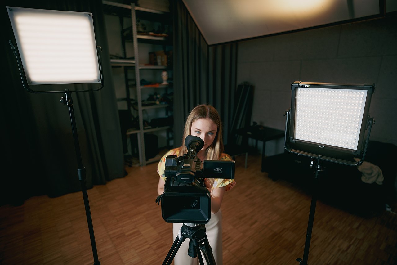 MZ_23_KRENN_v1-145 Eine Jugendliche steht zwischen zwei LED-Flächenleuchten und schaut durch den Sucher einer Panasonic-Videokamera.