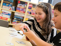 Zwei Kinder sitzen um einem Spieltisch und lachen bei einem Spiel, in dem Sie Begriffe auf Karten zeichnen.