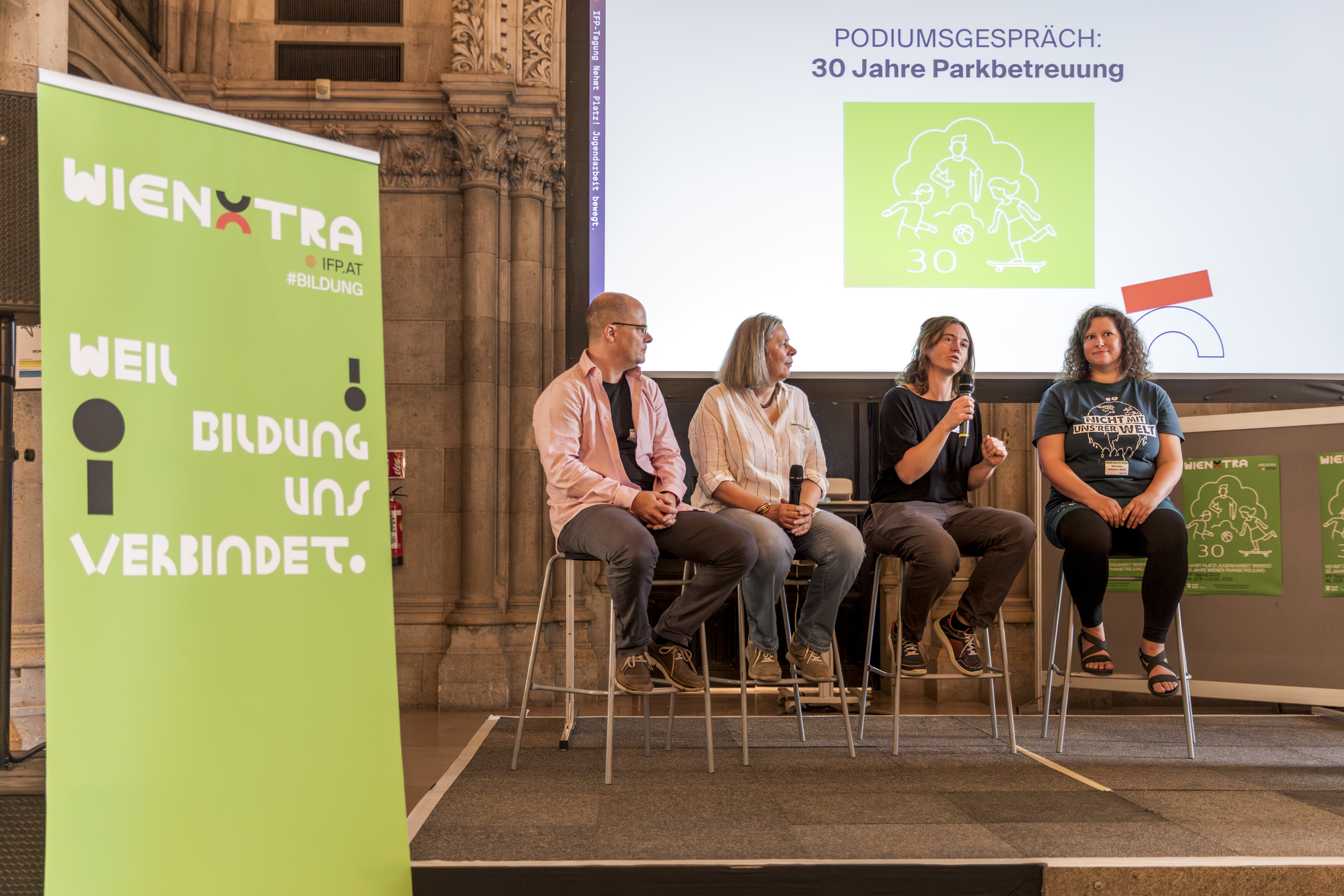 WIENXTRA-IFP-Tagung 2023: Nehmt Platz! Jugendarbeit bewegt. Vier Personen sitzen auf Hochstühlen auf einer Bühne und schauen sich an. Im Hintergrund ist eine Leinwand mit Projektion zu sehen. Auf der linken Seite steht ein Aufsteller.