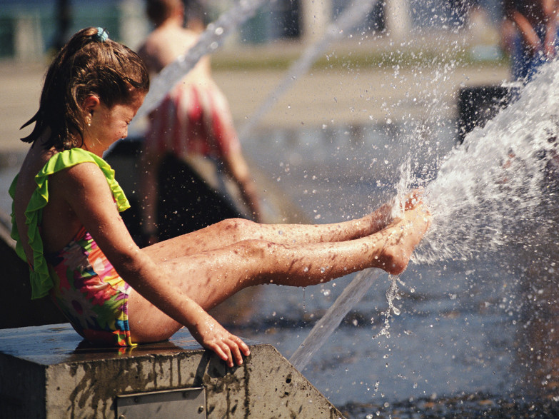 Wasserplanschen Maedchen sitzt am Wasserrand und spritzt mit den Fuessen Wasser hoch.