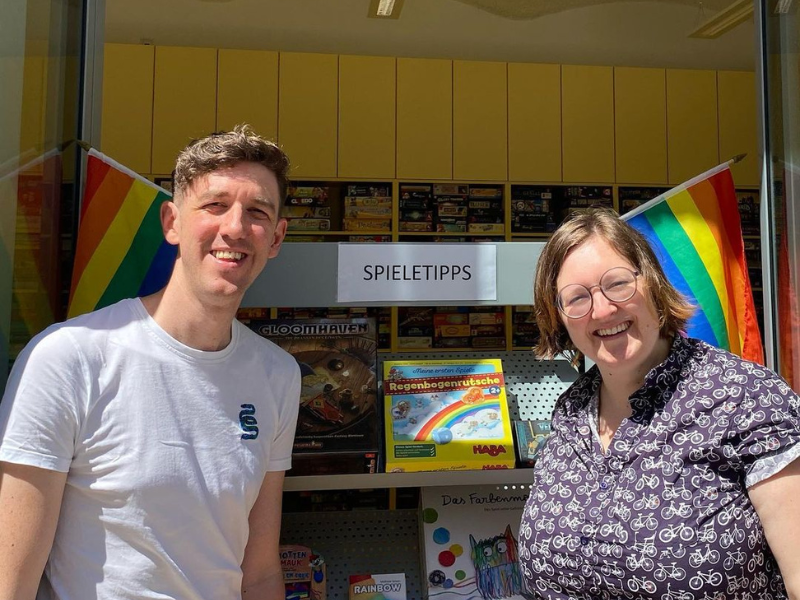 Vielfalt in der Wienxtra Spielebox Zwei Mitarbeiter_innen der Spielebox vor Spielen mit Regenbogenfahnen daneben