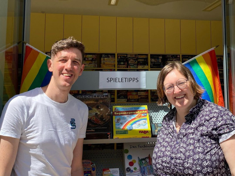 Zwei Mitarbeiter_innen der Spielebox vor Spielen mit Regenbogenfahnen  daneben