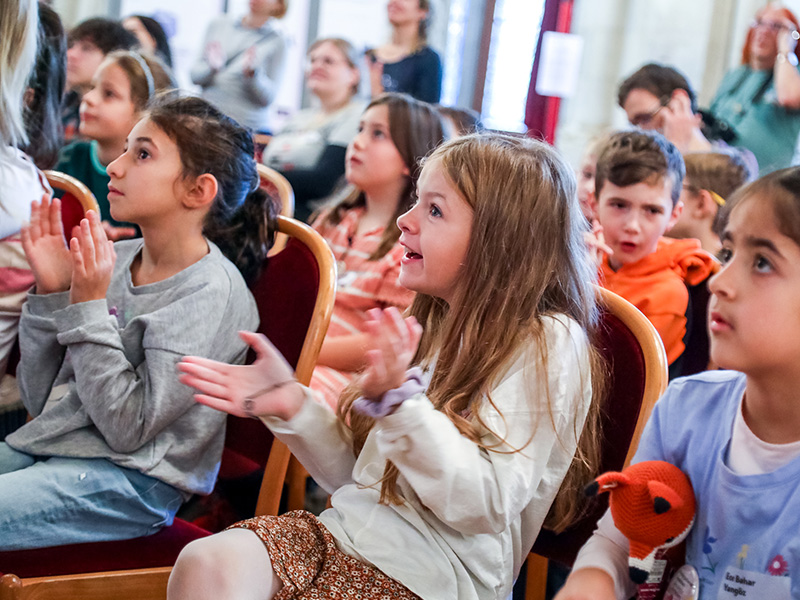 Drei Mädchen im Vordergrund und weitere Kinder in den hinteren Reihen applaudieren. 