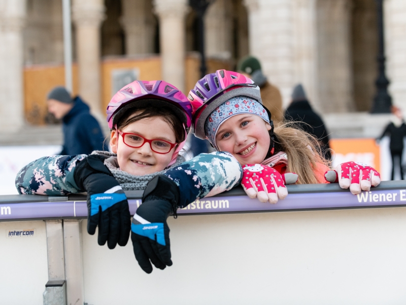SFS Zwei Mädchen sind am Eilaufplatz und lehnen sich an der Absperrung an, sie tragen Helme und Handschuhe. Ein Mädchen hat eine rote Brille, das andere lockige Haare.