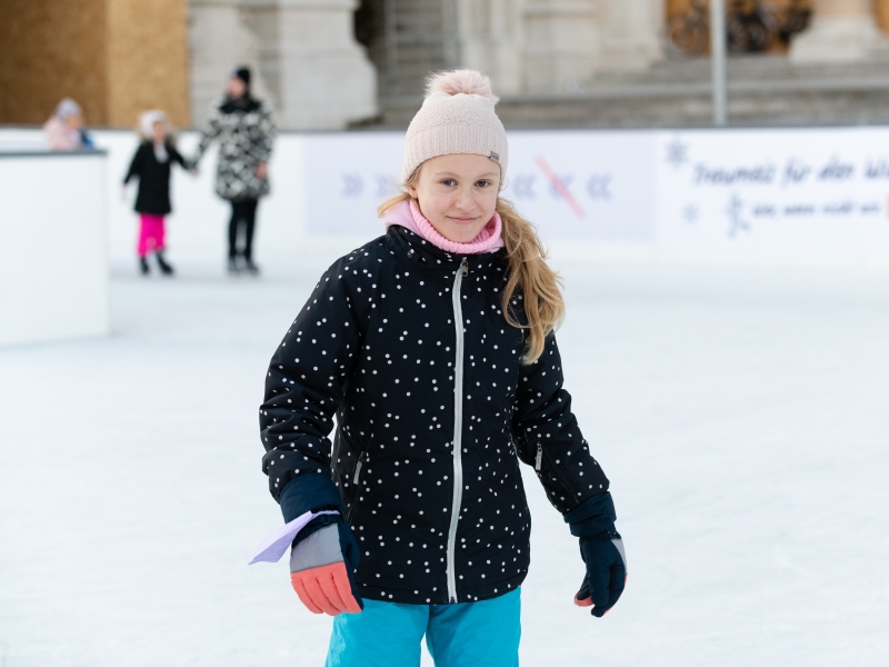 SFS Eine Teenagerin läuft Schlittschuh. Sie blickt freundlich in die Kamera. Sie hat blonde, lockige, lange Haare und trägt eine rosa Haube mit einem großen rosa Bommel. Ihre Jacke ist schwarz mit vielen kleinen rosa Punkten. Sie trägt eine blaue Skihose und grau-rosa Handschuhe.