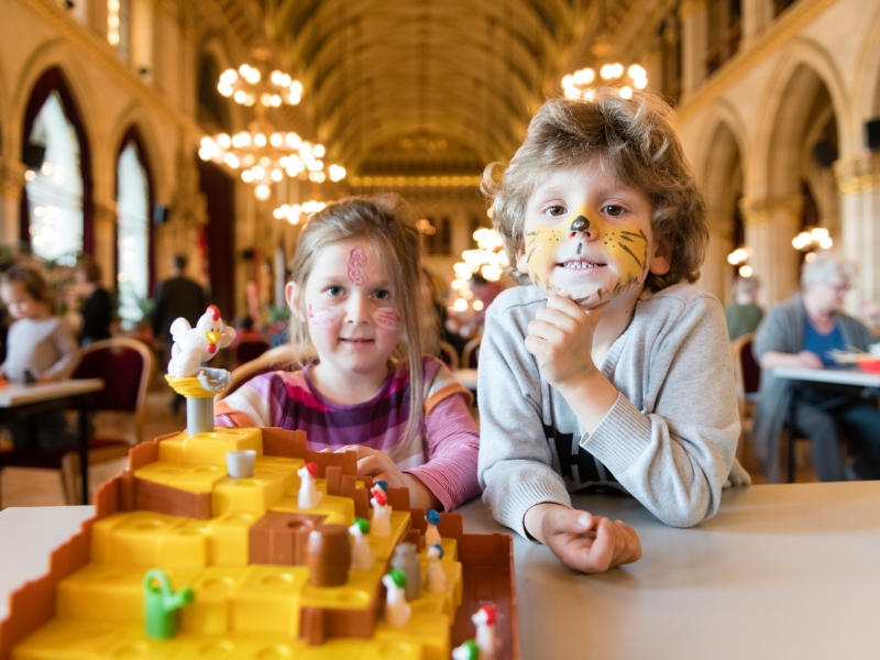 SFS Ein Mädchen und ein Bub sind bei einem Spielefest im Rathaus und sitzen an einem Spieletisch vor einem Kinderspiel mit einem Hühnchen. Sie haben geschminkte Gesichter. Der Bub ist ein Tiger und das Mädchen hat ein buntes Muster im Gesicht.