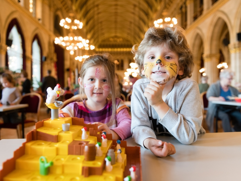 Ein Mädchen und ein Bub sind bei einem Spielefest im Rathaus und sitzen an einem Spieletisch vor einem Kinderspiel mit einem Hühnchen. Sie haben geschminkte Gesichter. Der Bub ist ein Tiger und das Mädchen hat ein buntes Muster im Gesicht.