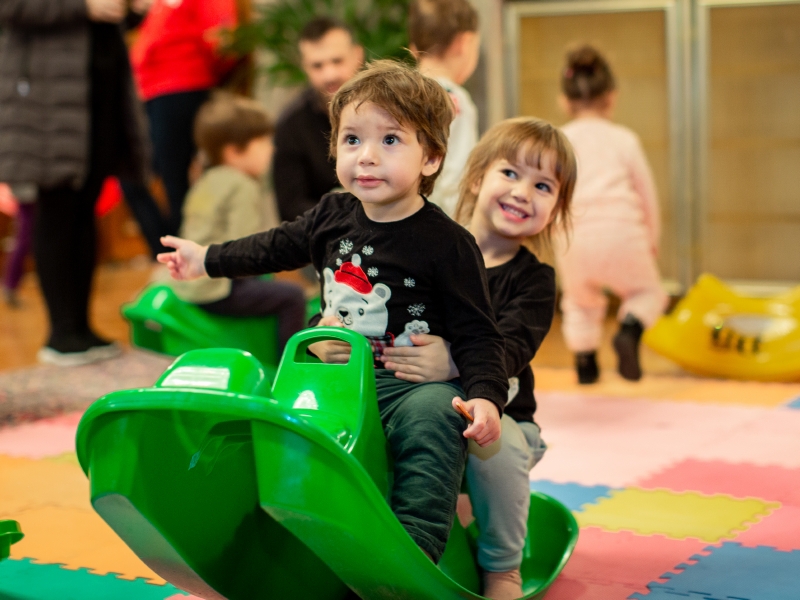 Zwei kleine Kinder sitzen auf einem grünen Schaukelbob. 