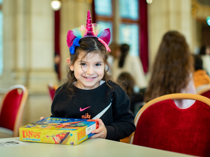 ST Ein Mädchen hat ein lustiges und glitzerndes Einhorn-Haarreif am Kopf und hält eine Brettspielschachtel in der Hand.